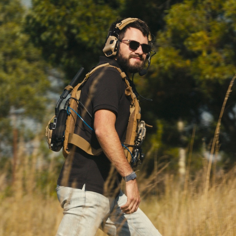 An operator walking through long grass in a headset and chest rig.