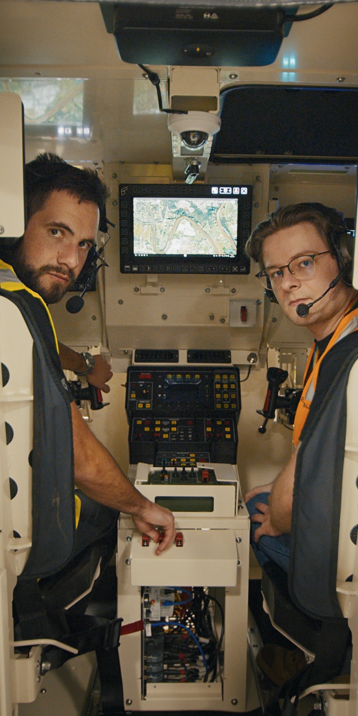 Two crew at a console inside a vehicle, working a mission display.