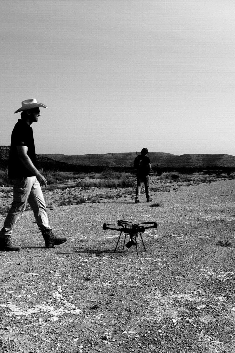 Two operators walking out across open scrub towards a quadcopter on the track.