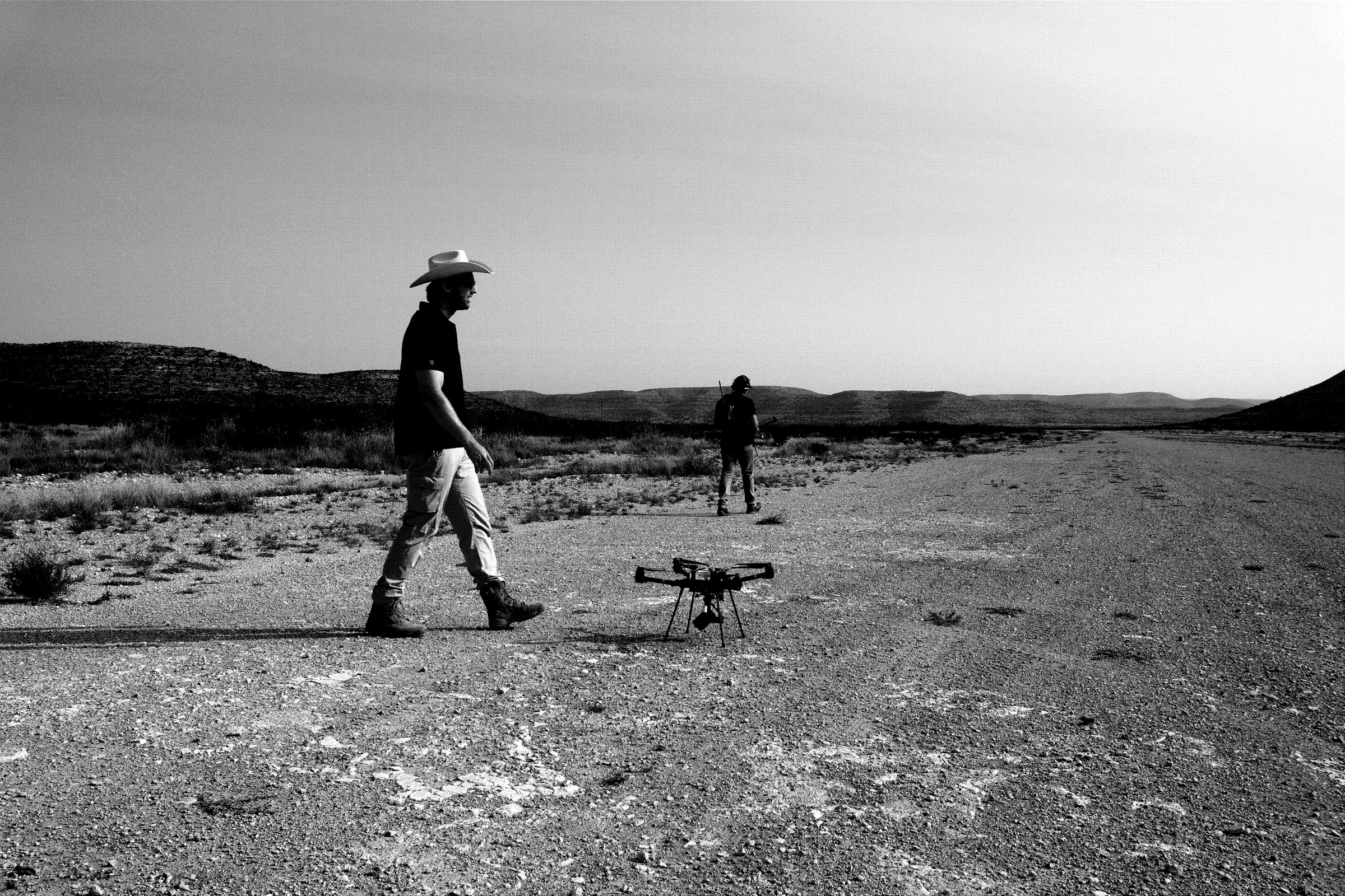 Two operators walking out across open scrub towards a quadcopter on the track.