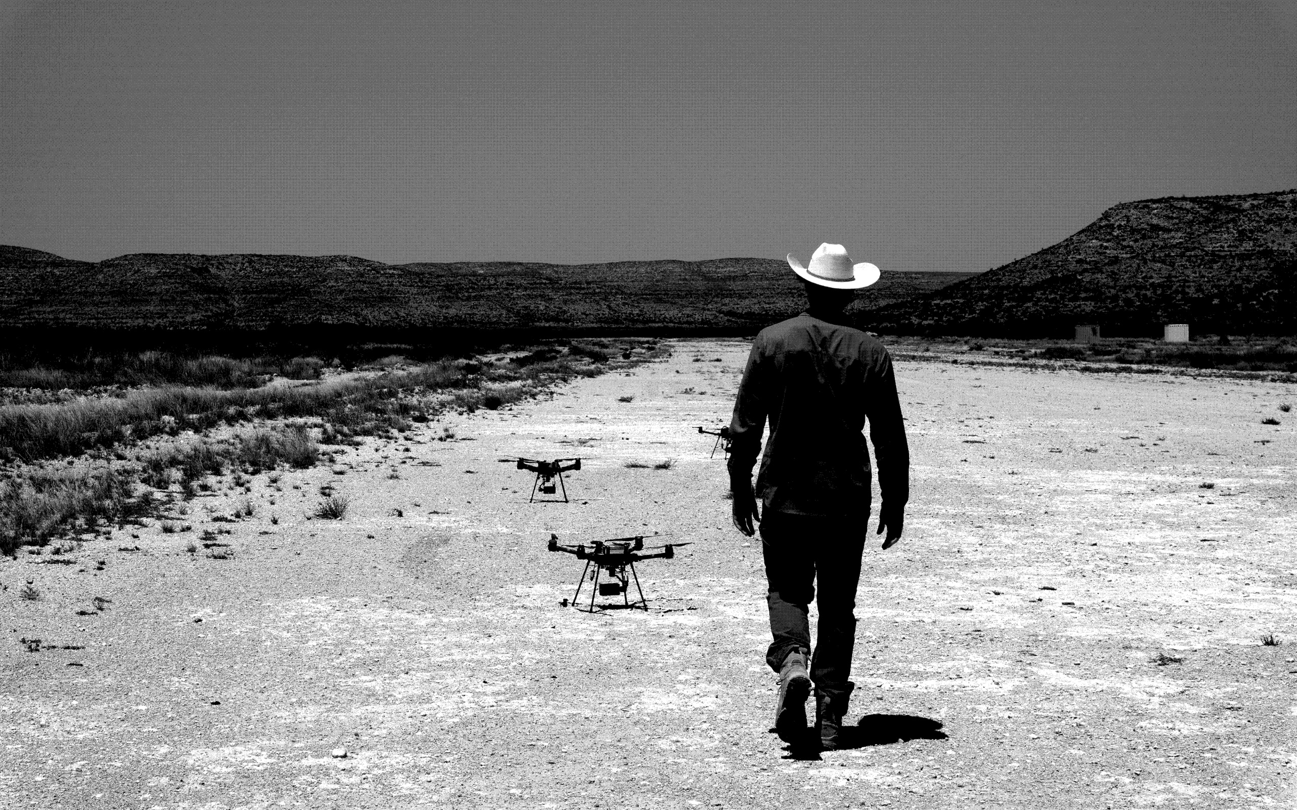 An operator walking away down a desert track towards two quadcopters on the ground.