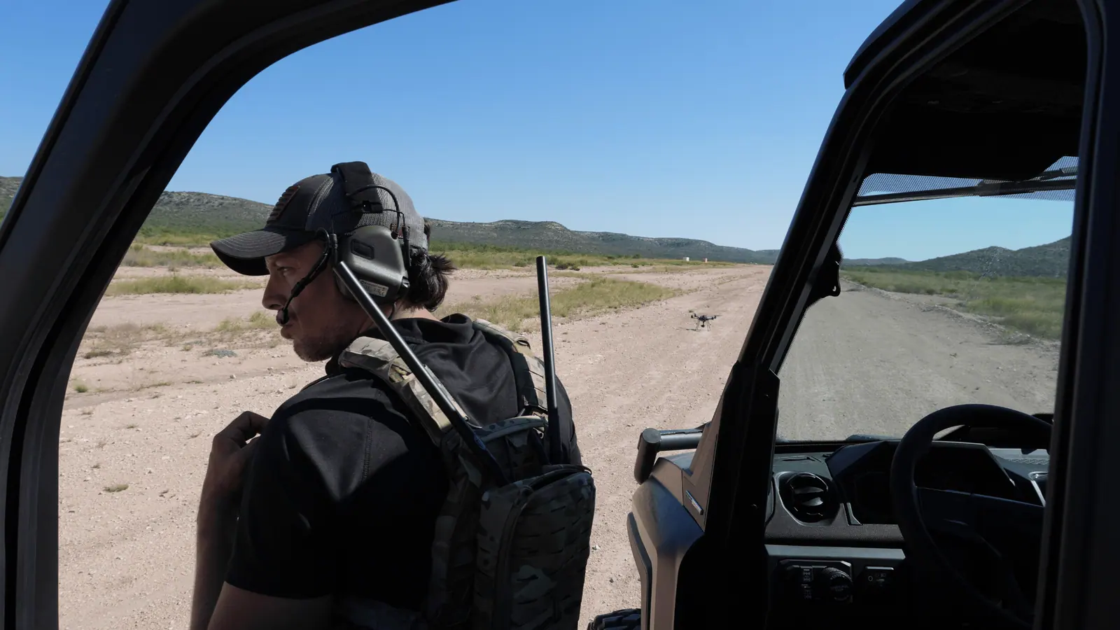 A Breaker operator in a headset stands beside a vehicle on a range, a drone flying over the track ahead.