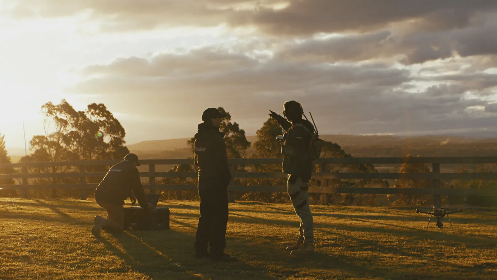 A Breaker team at sunset beside a fence line, one operator pointing out across the field, a drone on the grass beside them.
