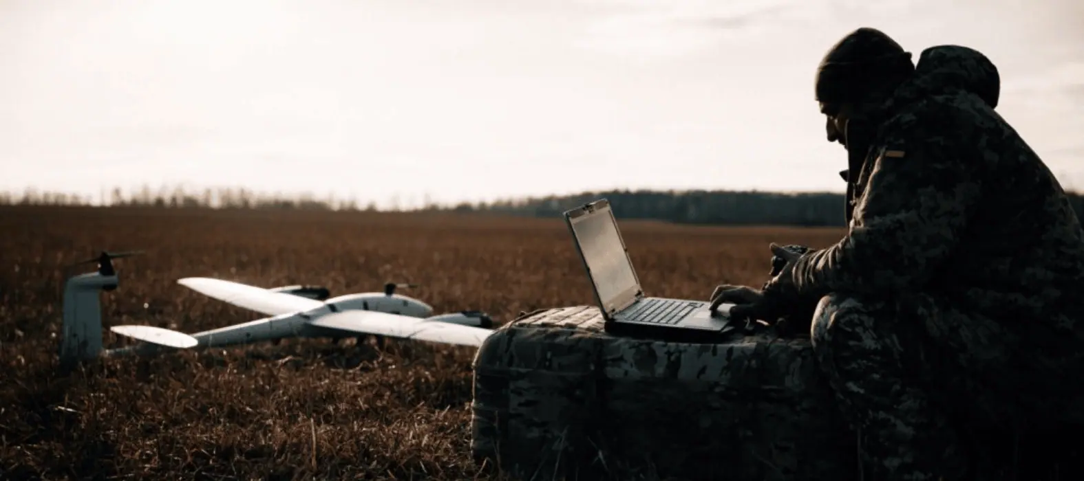An operator at a laptop in a field, a fixed-wing aircraft on the ground beside them.
