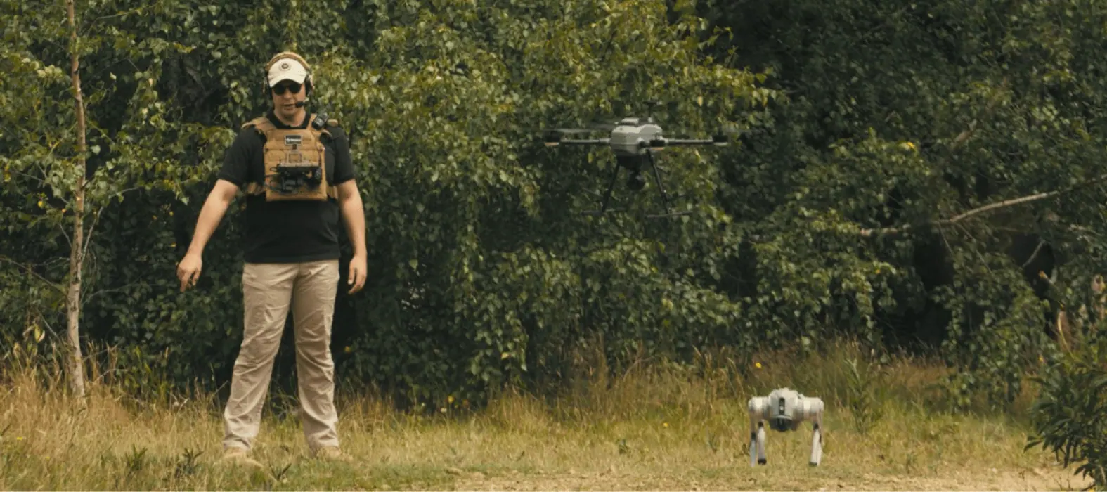 An operator in open ground with a fixed-wing aircraft overhead and a legged robot beside them.