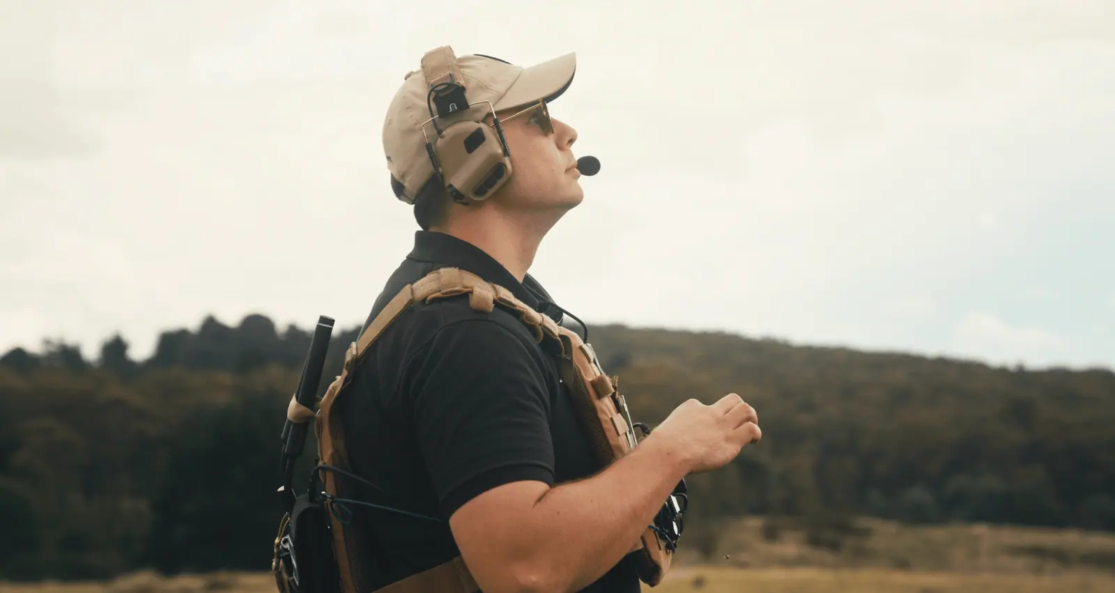 An operator in a plate carrier and headset looks up at the sky in open country.